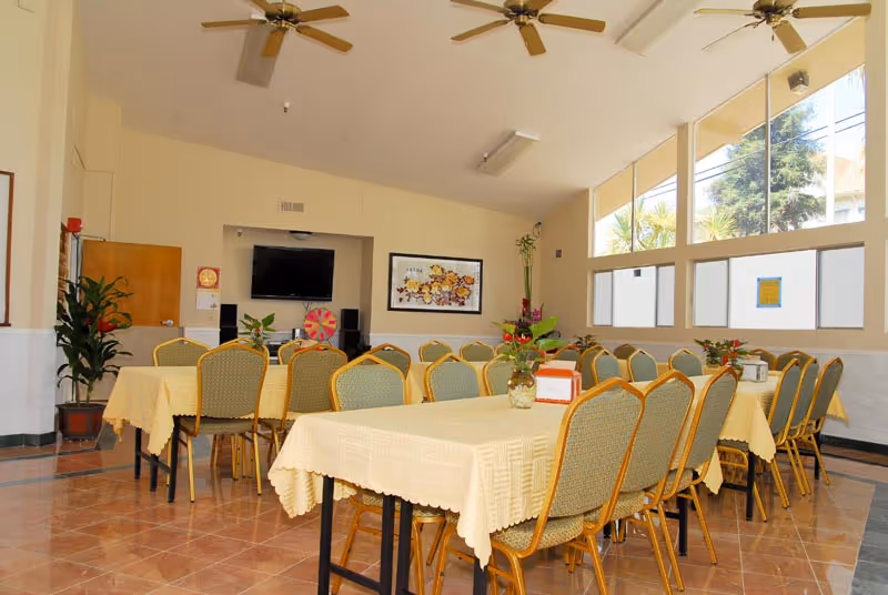 Bright dining room with long tables covered in yellow tablecloths, rows of chairs, large windows and ceiling fans.