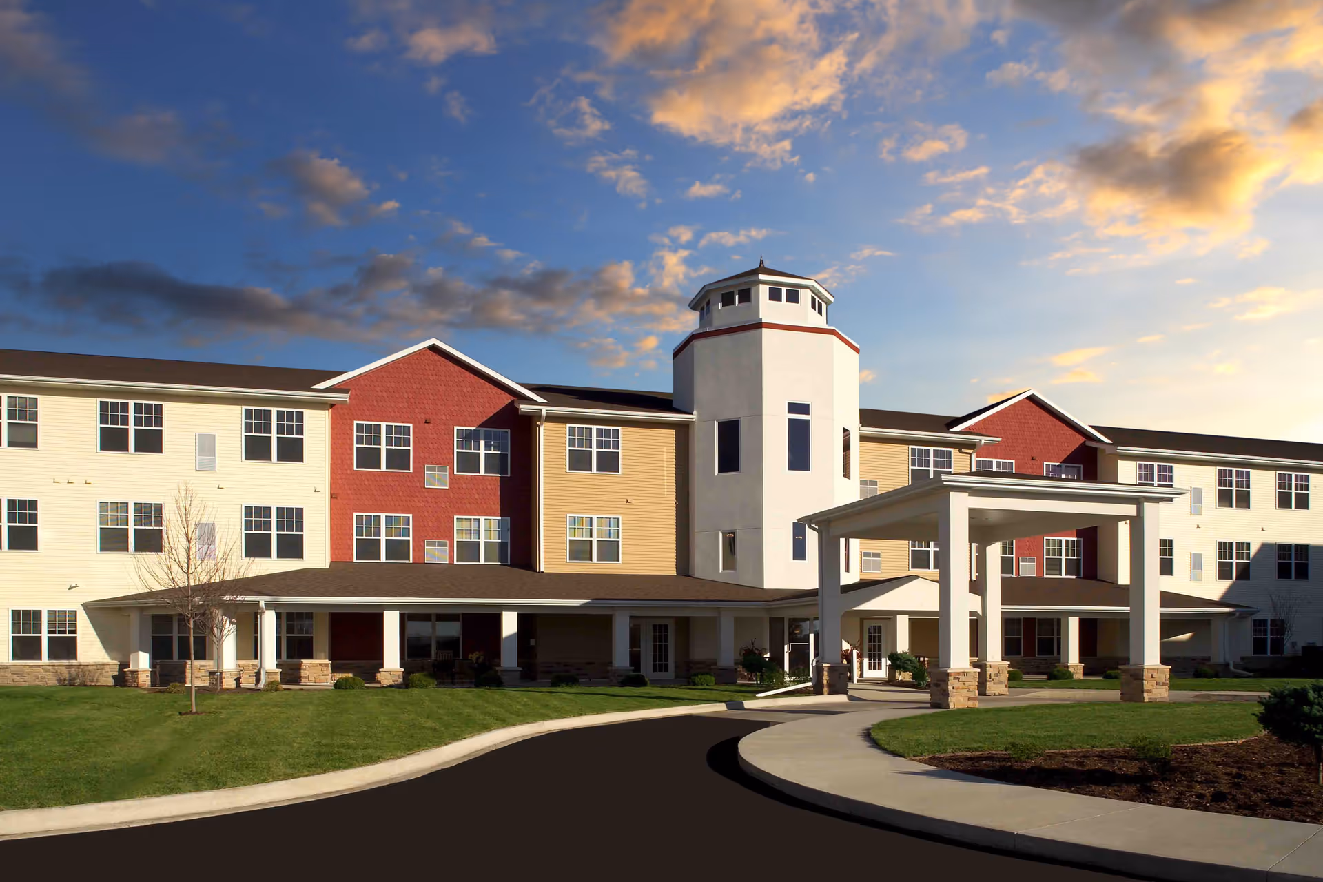 Exterior view of a multi-story senior living facility building with a covered entrance, manicured lawn, and a partly cloudy sky at sunset.