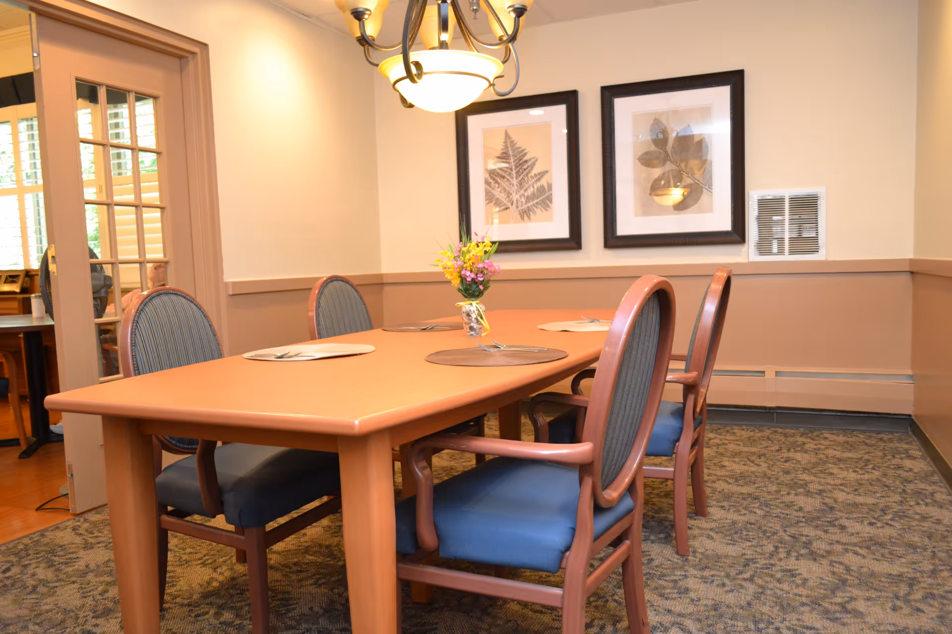 A dining room with a wooden table set for four, featuring blue cushioned chairs. A small vase with flowers is placed in the center of the table. The room has beige walls with two framed botanical prints hanging on one wall and a chandelier overhead. A door with glass panels leads to another room.