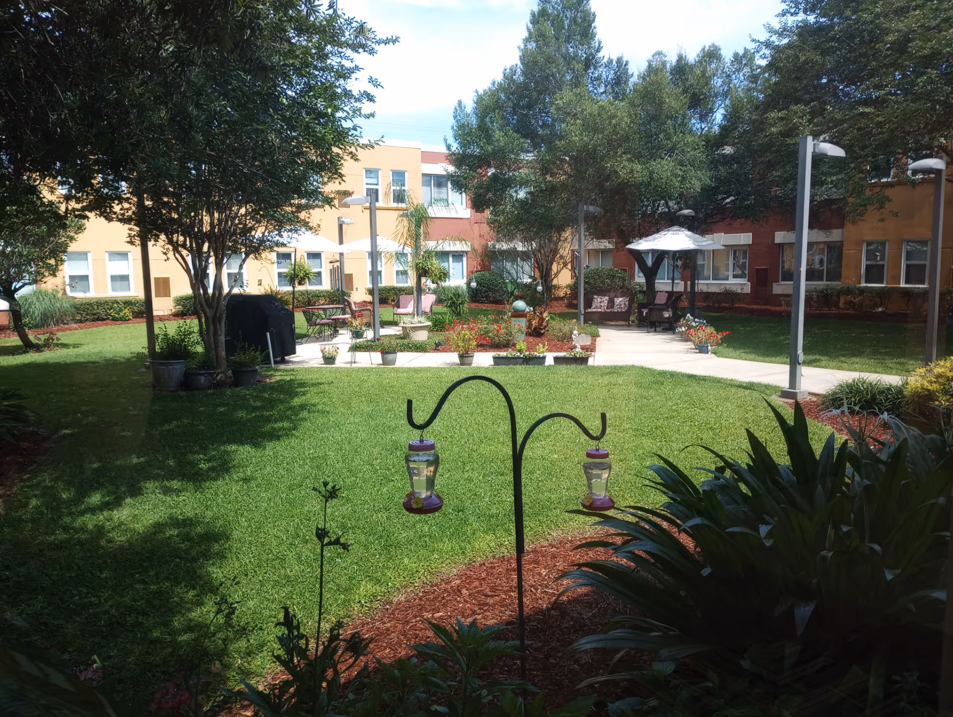 A sunny courtyard garden area with green grass, trees, potted plants, and a paved walkway. There are benches, a gazebo with a white canopy, and a building with multiple windows surrounding the garden.