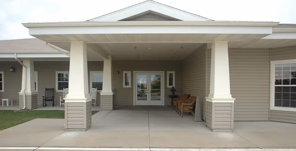 Covered entrance area of a building with beige siding, white pillars, and a double glass door. There are chairs and a small table on either side of the entrance, and a concrete driveway in front.