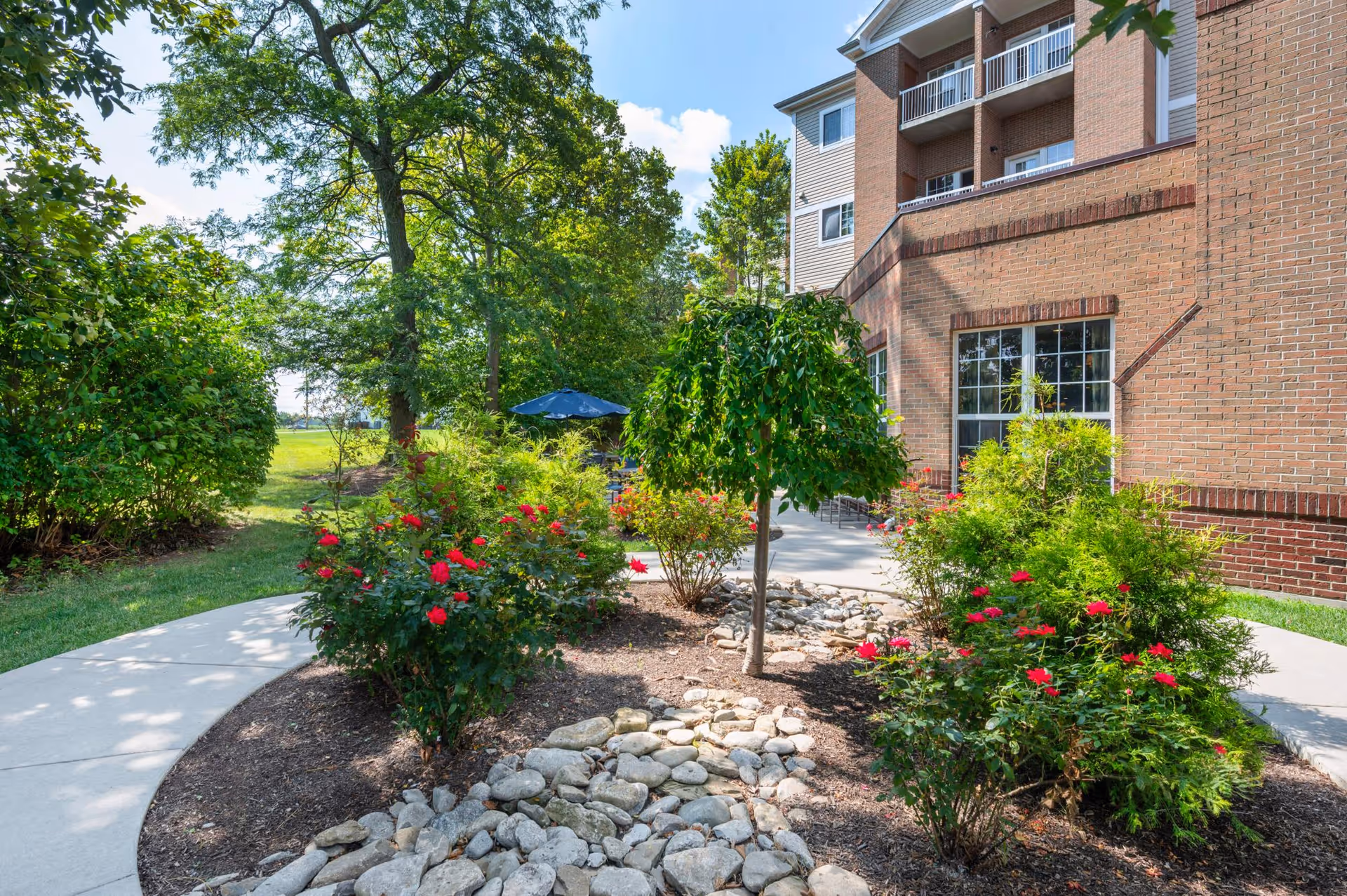 Outdoor garden area at Barrington of West Chester featuring a curved concrete walkway, landscaped flower beds with red flowers and green shrubs, a small tree, and a brick building with balconies in the background under a partly cloudy sky.
