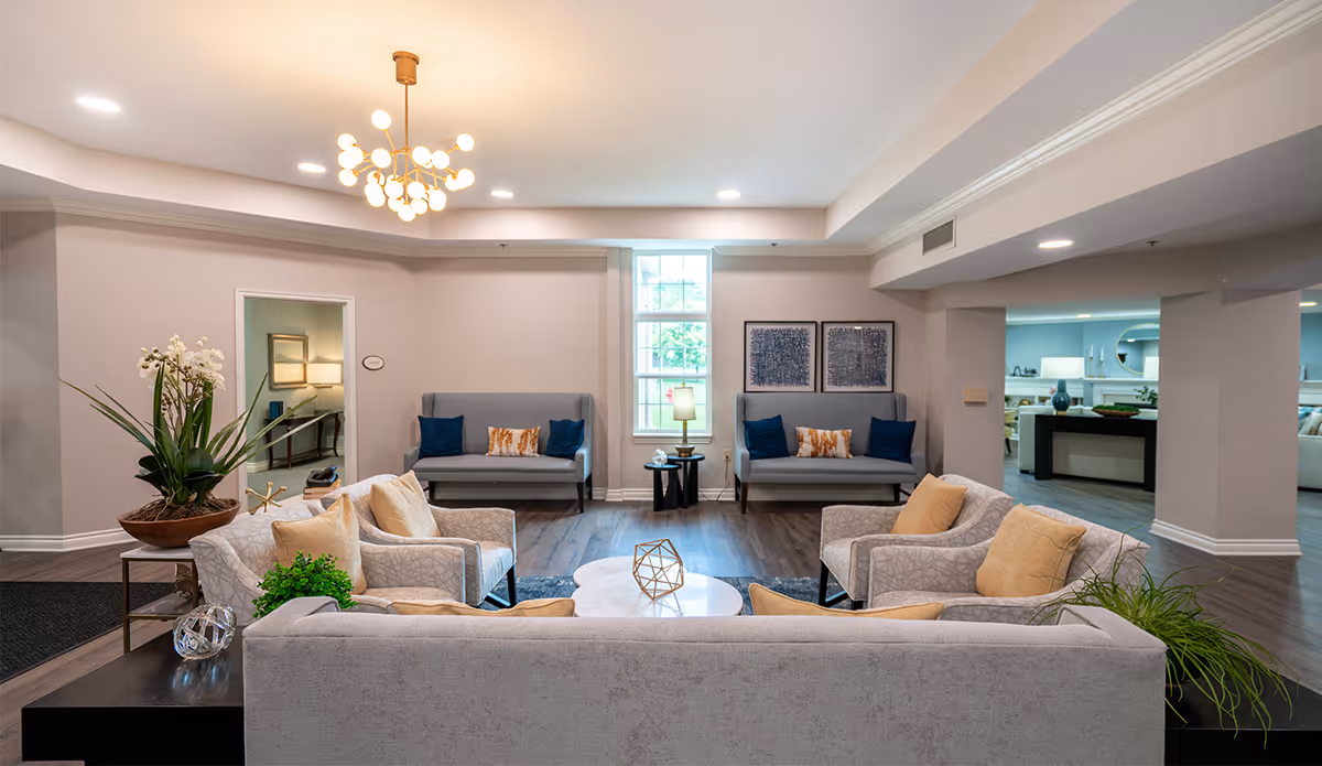 A bright and spacious living room area in a senior living facility with modern decor. The room features multiple gray sofas and armchairs with beige and blue cushions arranged around a white circular coffee table. There are decorative plants on side tables, a large window letting in natural light, two framed artworks on the wall, and a contemporary chandelier hanging from the ceiling.