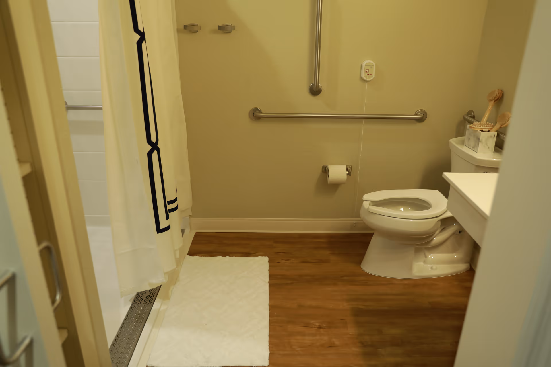 A bathroom with a white toilet, a white shower curtain with black geometric patterns, a white bath mat on a wooden floor, grab bars on the wall, and a small countertop with a tissue box and wooden brushes.