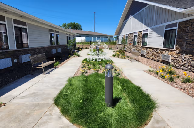 Outdoor courtyard area between two single-story buildings with stone and siding exteriors. The courtyard has a concrete walkway, green grass, flower beds with yellow flowers, benches along the walkway, and a decorative white metal bench shaped like a carriage at the far end.