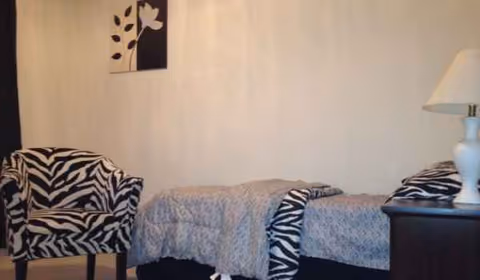 A bedroom in a senior living facility with zebra-pattern chairs, a bed, and a lamp.