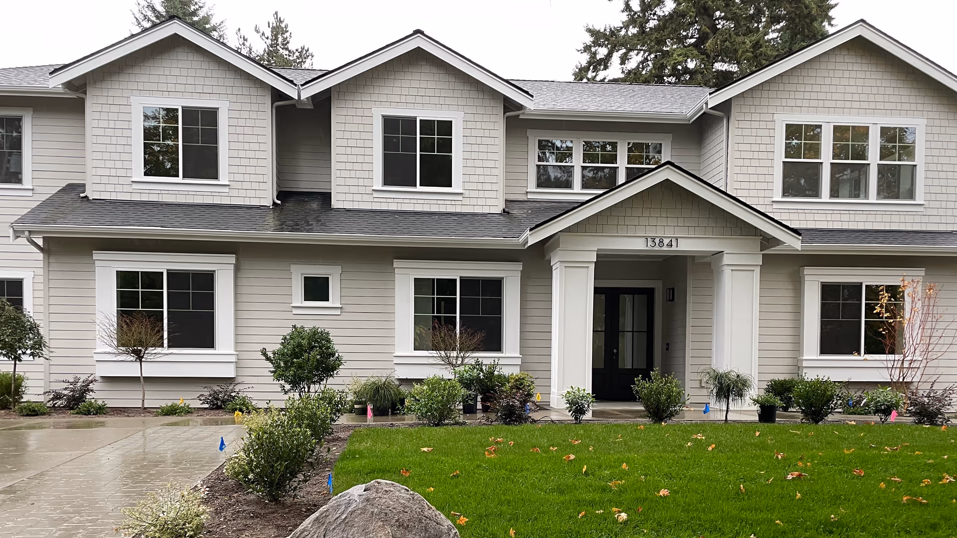 Front exterior view of a two-story senior care facility building with beige siding, white trim, multiple windows, a covered entrance with columns, and a well-maintained lawn with shrubs and small trees.