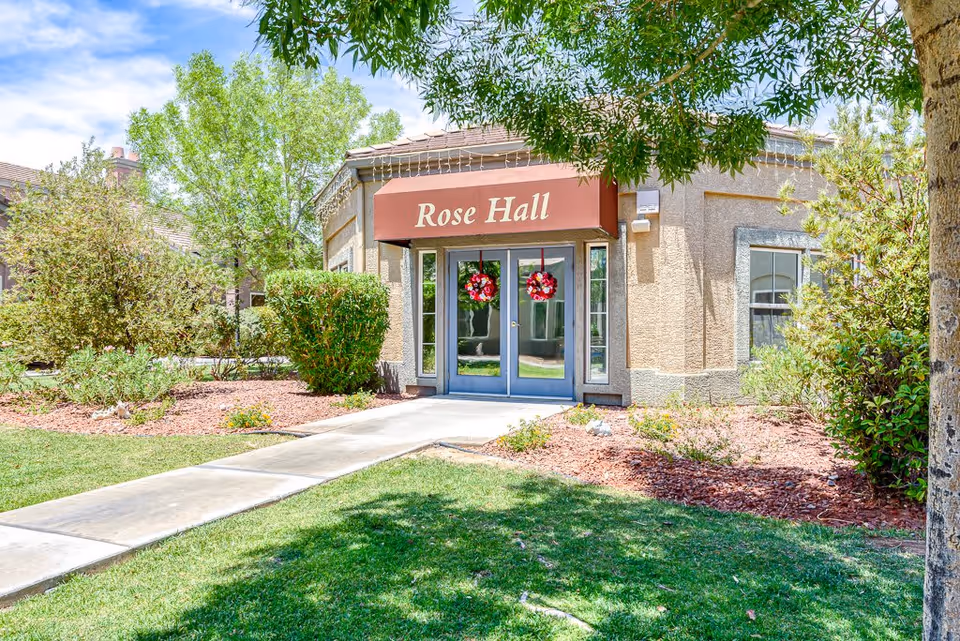 Exterior view of a building named Rose Hall at Cottages at Green Valley, featuring double glass doors decorated with red wreaths, surrounded by landscaped greenery and a concrete walkway leading to the entrance.