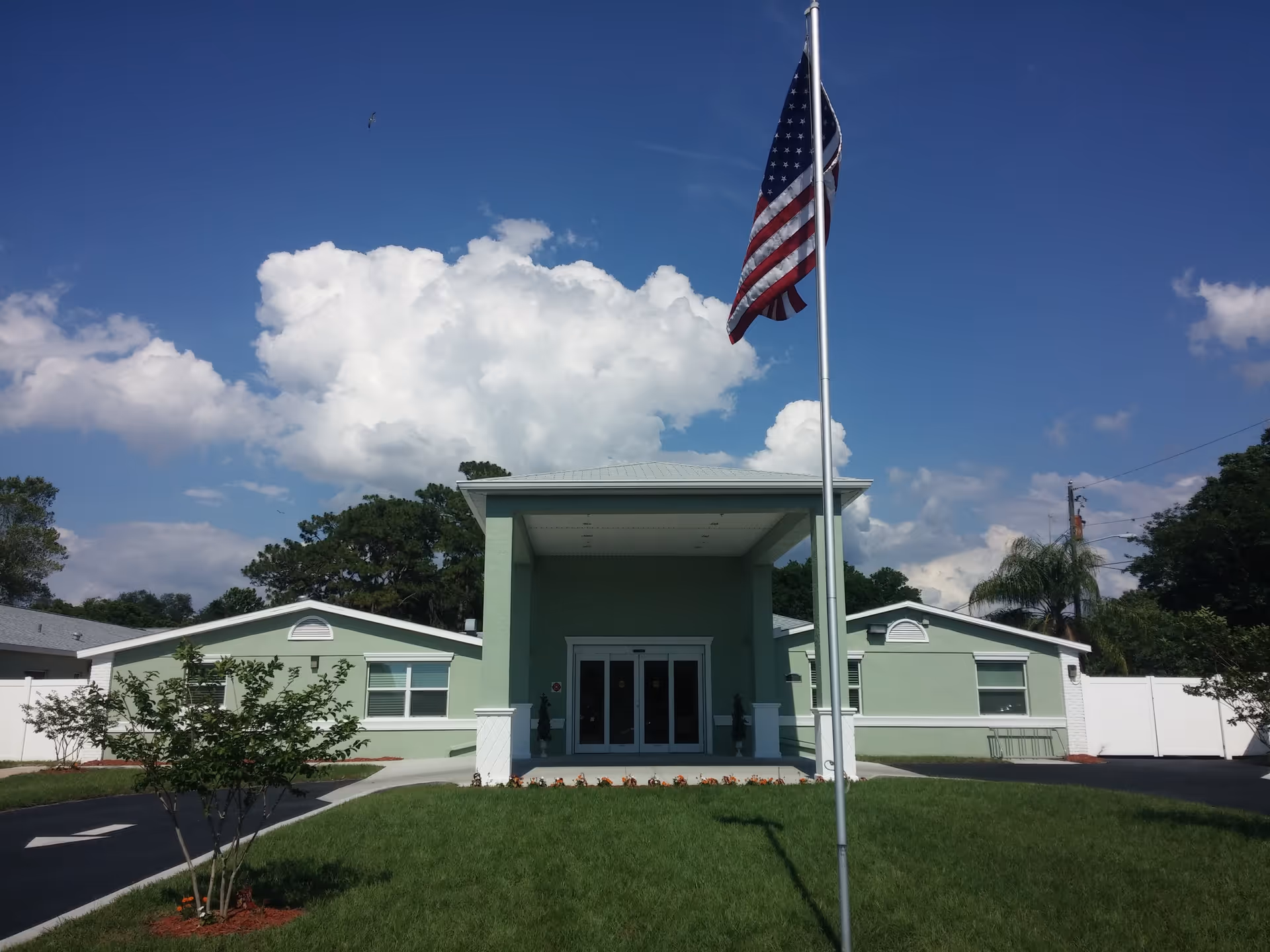 Front exterior view of a single-story green building with a covered entrance, an American flag on a flagpole in the foreground, and a well-maintained lawn and trees under a partly cloudy blue sky.