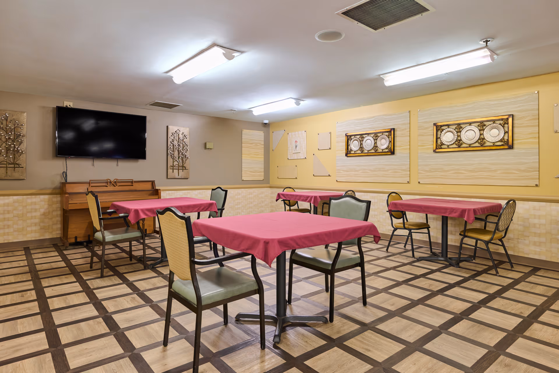 Empty communal dining/activity room with tables covered in red cloths, chairs, a wall-mounted TV, and decorative wall art.