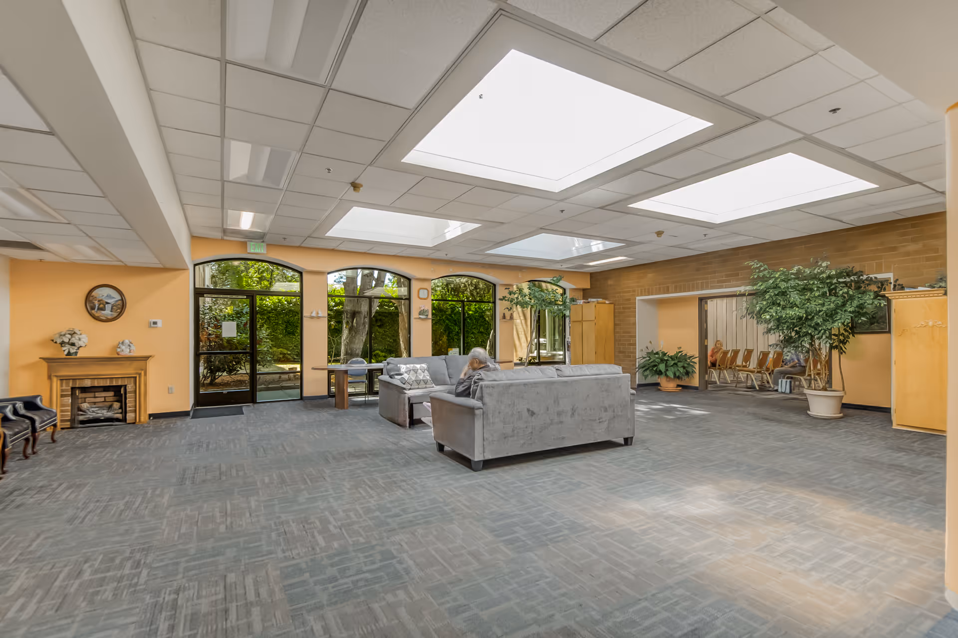 A spacious common area with large skylights in the ceiling allowing natural light to fill the room. The room has light orange walls, large windows and glass doors looking out to greenery. There are two gray sofas arranged around a small table, a fireplace on the left wall, several potted plants, and a few chairs lined up in an adjacent room visible through an open doorway.