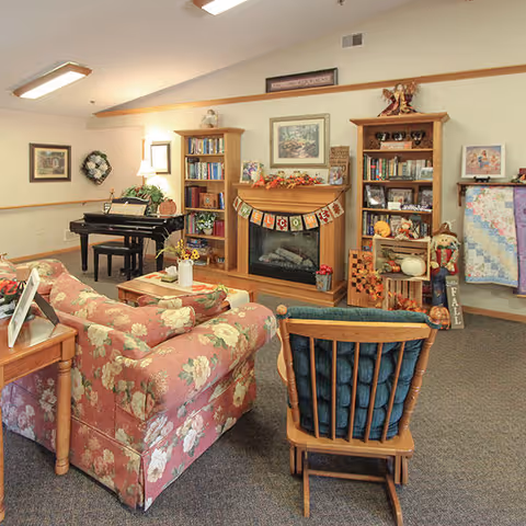 Cozy senior living common room with floral sofas, a wooden armchair, fireplace flanked by bookshelves, and a piano in the background.