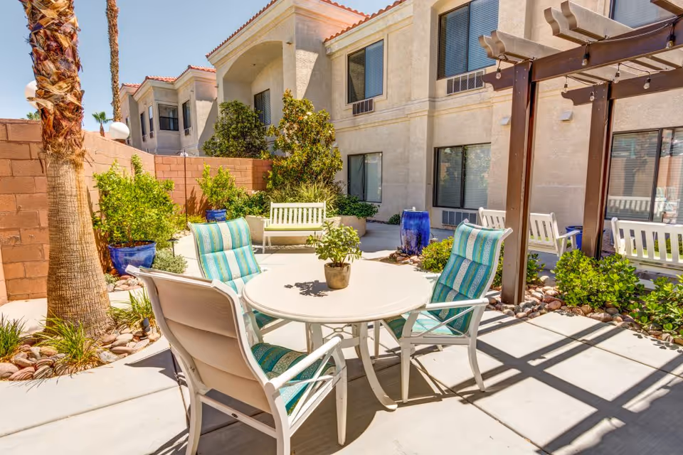 Outdoor patio area with a round white table surrounded by four cushioned chairs with blue and green striped upholstery. There are potted plants, a palm tree, and a wooden pergola casting shadows on the concrete floor. Beige building walls with windows and white benches are visible in the background under a clear blue sky.