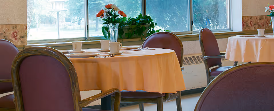 A dining area with round tables covered in light orange tablecloths, set with white cups and plates. Purple cushioned chairs with wooden armrests surround the tables. Large windows in the background let in natural light and show greenery outside.