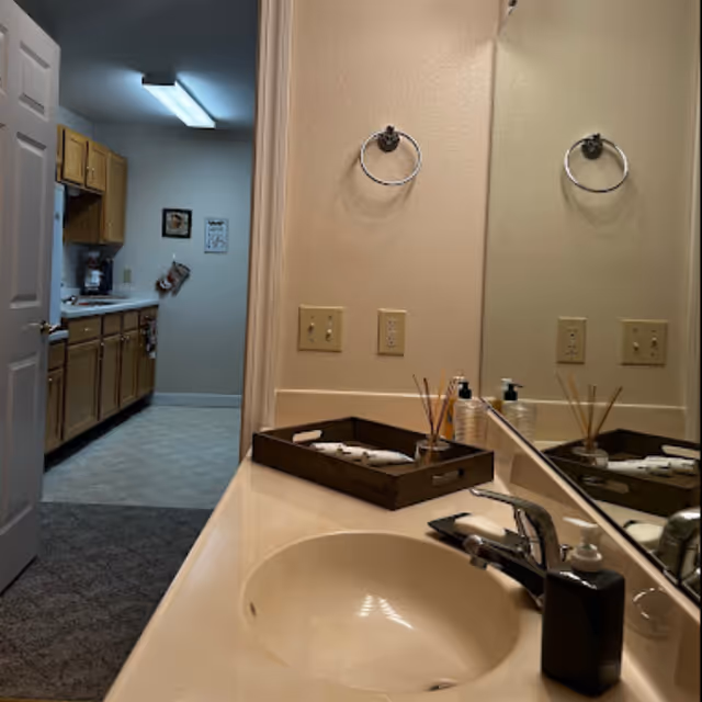 A bathroom sink countertop with a large mirror above it. On the countertop, there is a black soap dispenser, a tray holding toiletries, and reed diffusers. The wall has two towel rings and electrical outlets. Through an open door, a kitchen with wooden cabinets and a coffee maker is visible.
