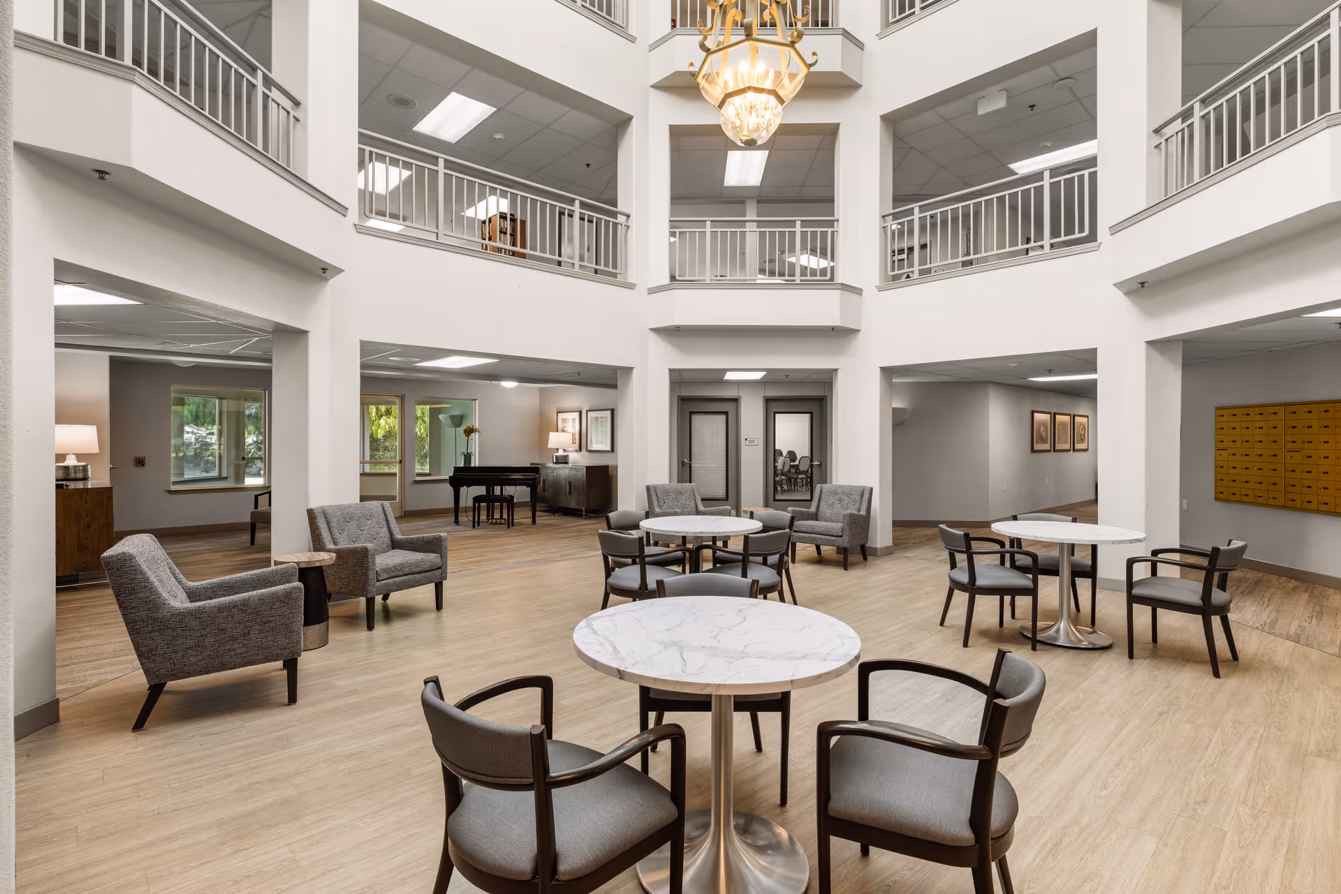 A spacious and well-lit common area in a senior living facility featuring round marble-top tables with chairs, several armchairs, a chandelier hanging from the ceiling, and a piano in the background. The area has light wood flooring, white walls, and a second-floor balcony overlooking the space.
