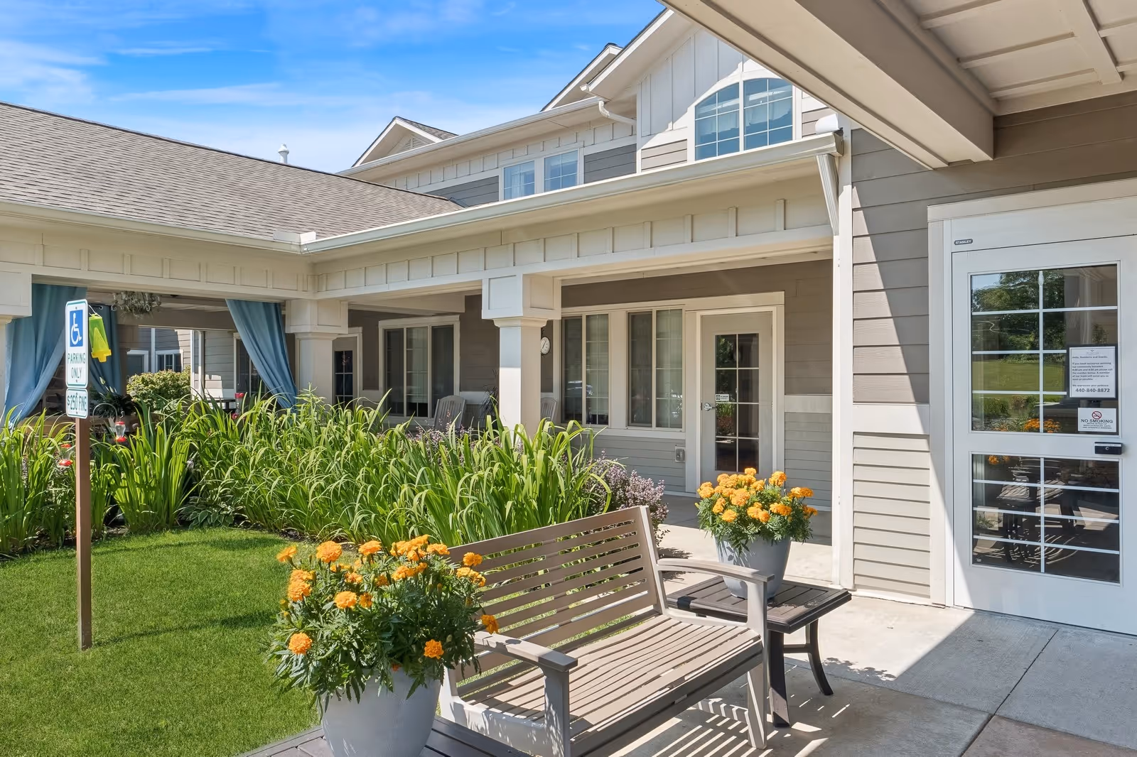 Outdoor seating area at The Enclave of Newell Creek featuring a wooden bench with armrests, two flower pots with yellow-orange flowers, green plants, and a building entrance with glass doors and windows under a covered porch.