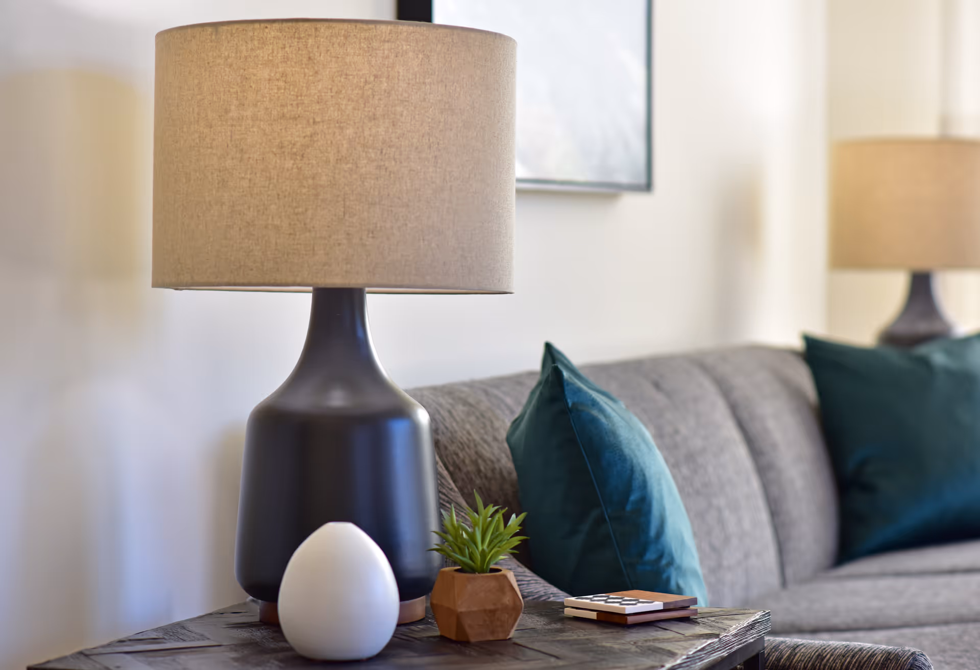 Side table with a lamp and decorative items next to a gray sofa with teal throw pillows in a living room.