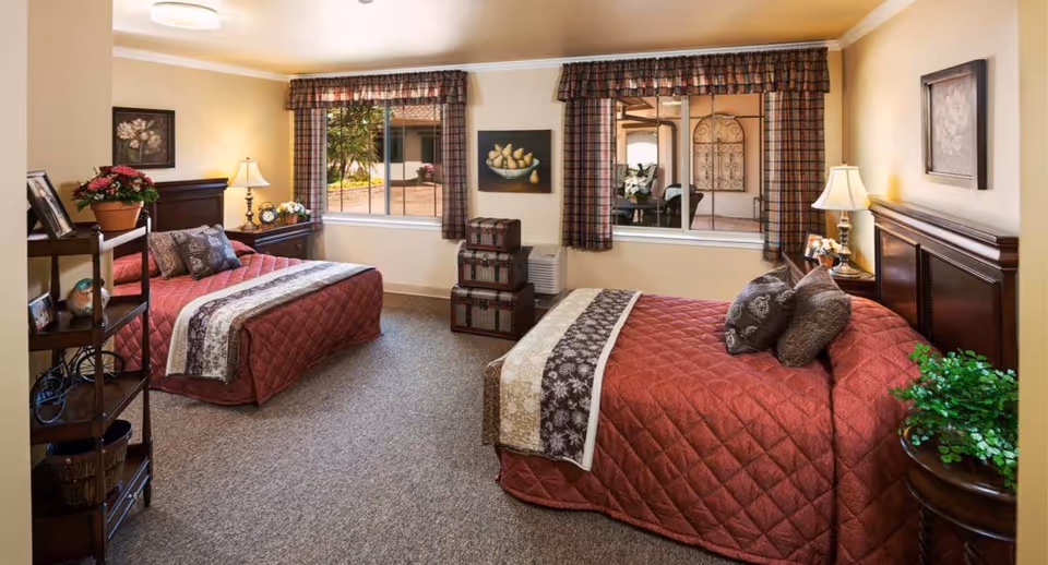 Sunlit twin bedroom with two red quilted beds, wooden headboards, bedside lamps, windows and decorative plants.