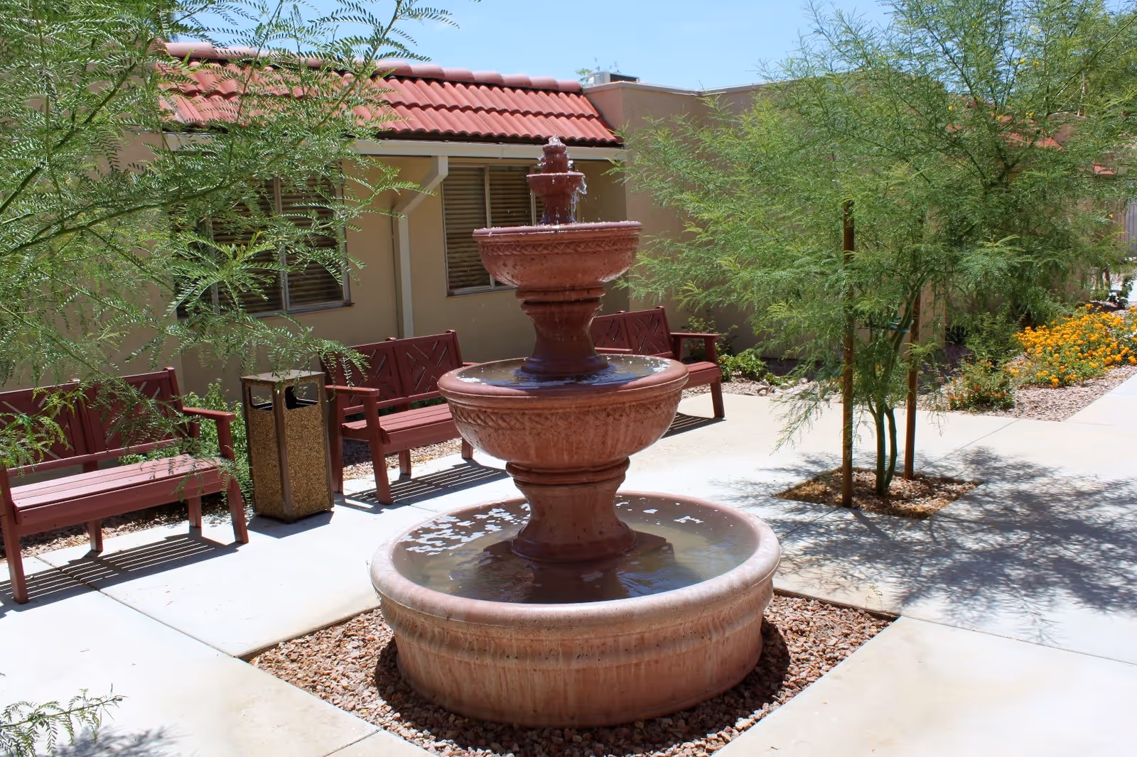 Outdoor courtyard area with a three-tiered stone water fountain in the center, surrounded by benches, small trees, and flowering plants. The building with a red tiled roof and windows is visible in the background under a clear blue sky.