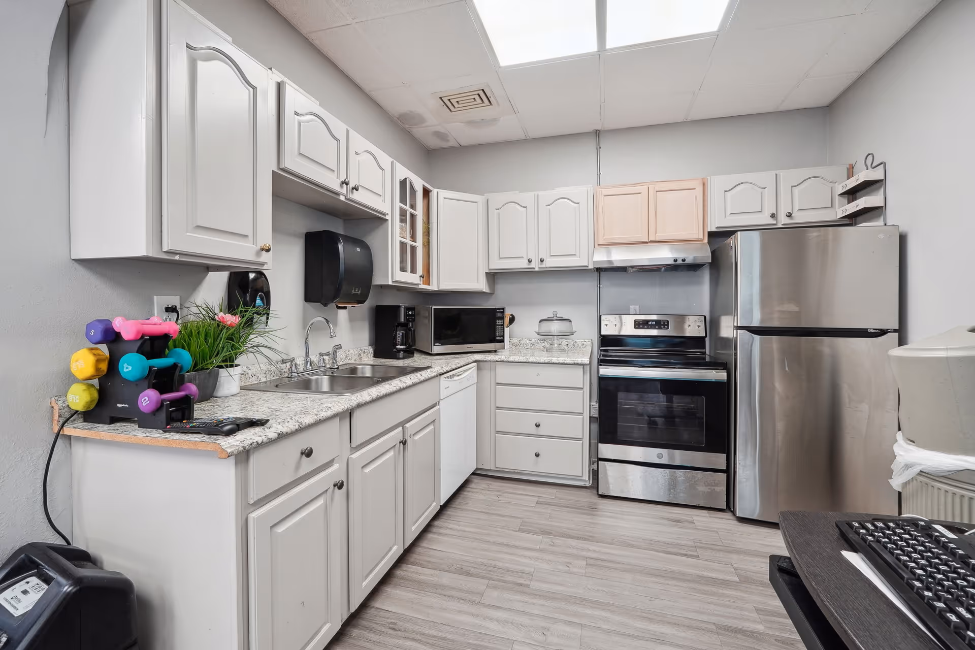 A clean and modern kitchen with white cabinets, granite countertops, a stainless steel refrigerator and oven, a microwave, a coffee maker, and a double sink. Colorful dumbbells and a small potted plant are placed on the countertop. The floor has light wood-style flooring and the ceiling has fluorescent lights.