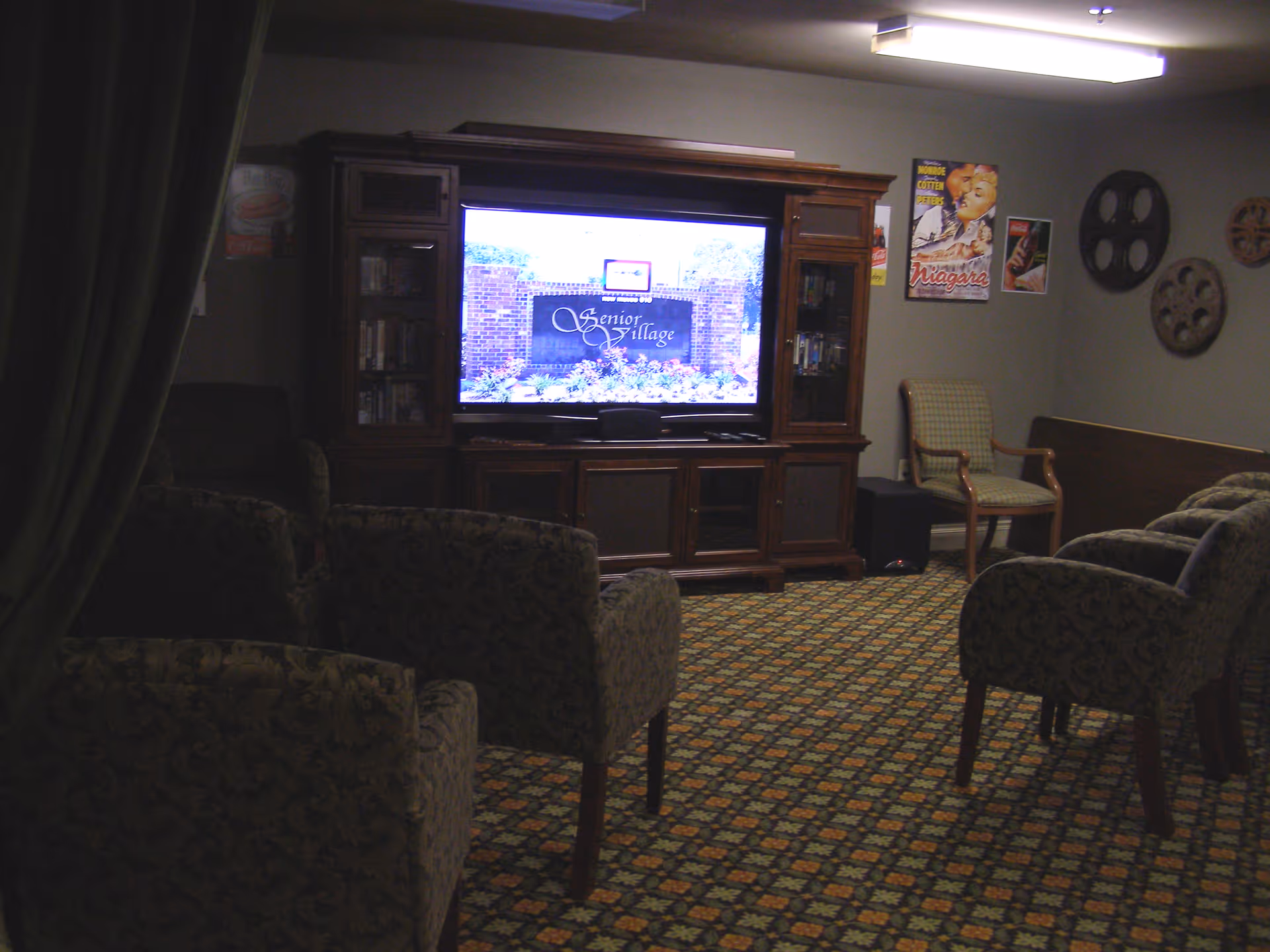 Dimly lit communal lounge with patterned armchairs facing a large TV in a wooden entertainment cabinet.