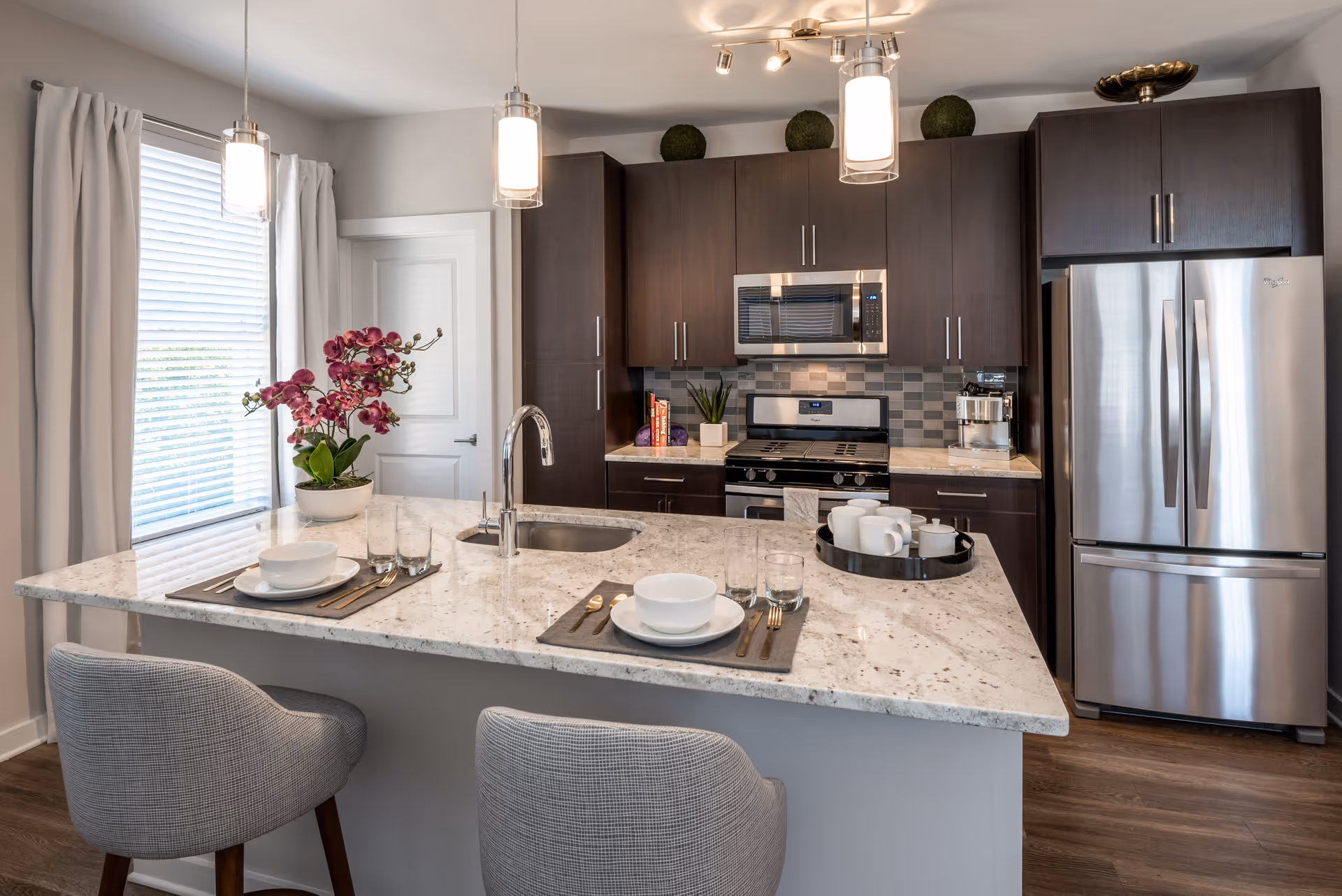 Modern kitchen with dark wood cabinets, stainless steel refrigerator and oven, a marble countertop island with two place settings, a sink, and a tray with cups. There are two pendant lights hanging above the island and a window with white curtains to the left.