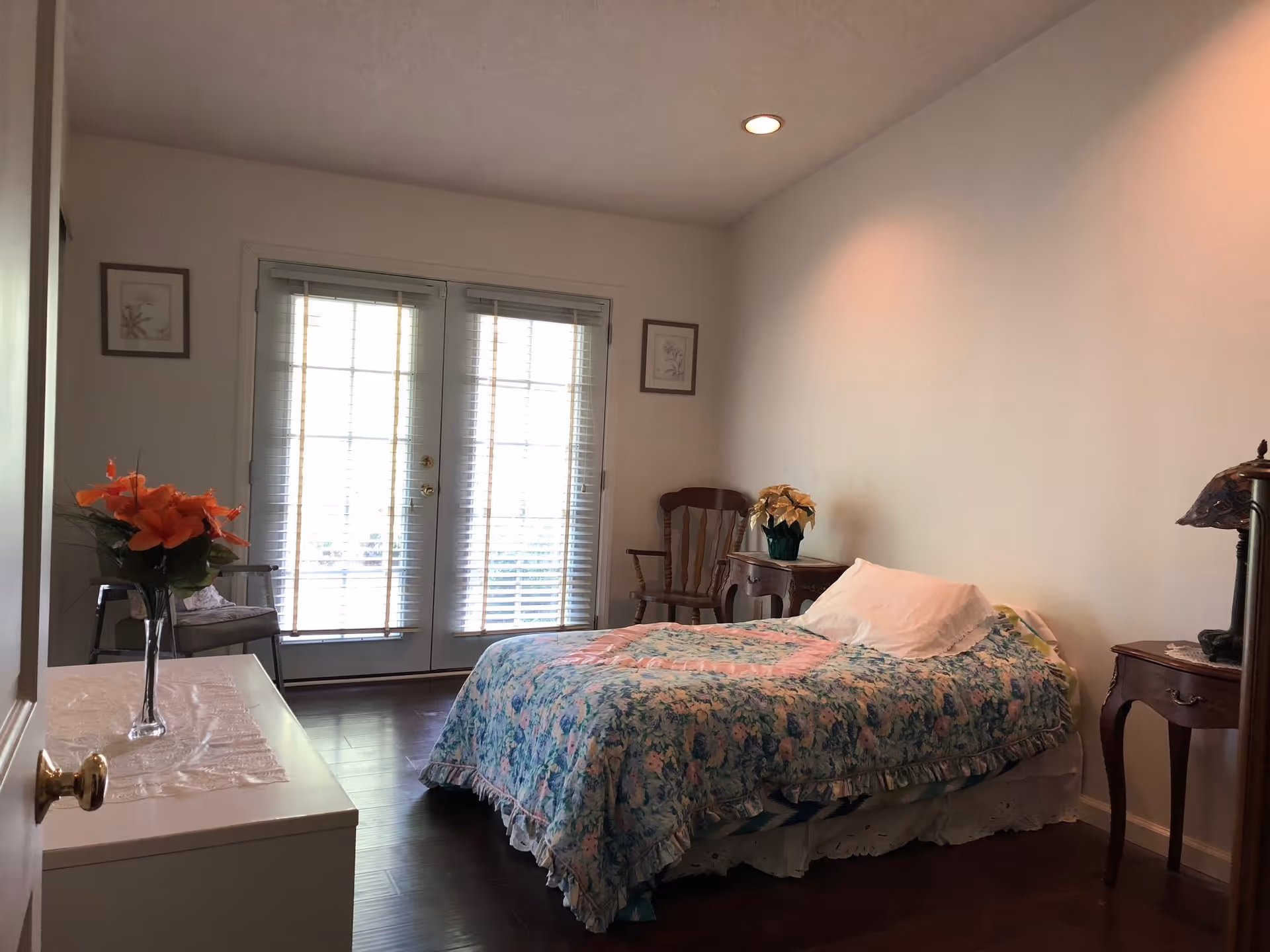 A bedroom with a single bed covered in a floral quilt, a white pillow, and a wooden chair next to a small wooden side table with a potted plant. There is a white dresser with a vase of orange flowers on it, two framed pictures on the walls, and French doors with blinds letting in natural light.