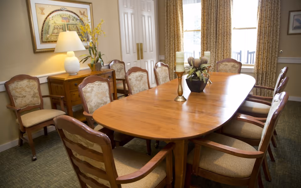 Wooden dining table surrounded by upholstered chairs in a bright dining room with a sideboard, lamp, and window curtains.