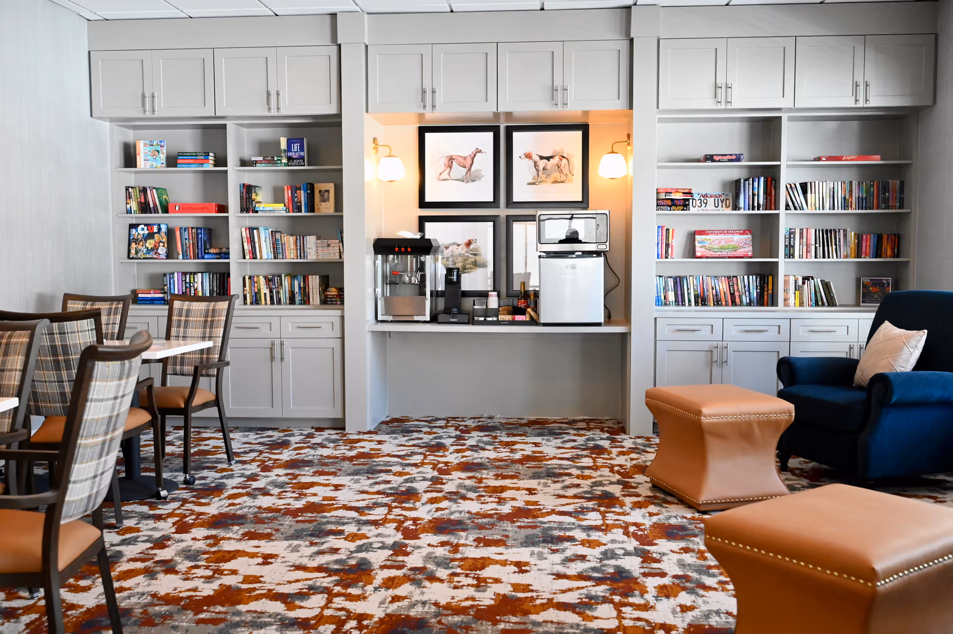 Bright common room with bookshelves, a coffee station, tables and chairs, and upholstered seating on a patterned carpet.