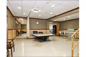 A rehabilitation room with a treatment table in the center, parallel bars for walking exercises on the right, a curtain divider, chairs along the left wall, and a clock on the far wall under ceiling lights.