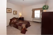 A bedroom with a single bed covered in a brown and beige checkered blanket. There is a wooden nightstand with a lamp and a small drawer next to the bed. Two framed pictures hang on the wall above the bed. A window with beige curtains is on the far wall, and a tall dark wooden dresser with a potted plant on top is on the right side of the room. The carpet is light beige.