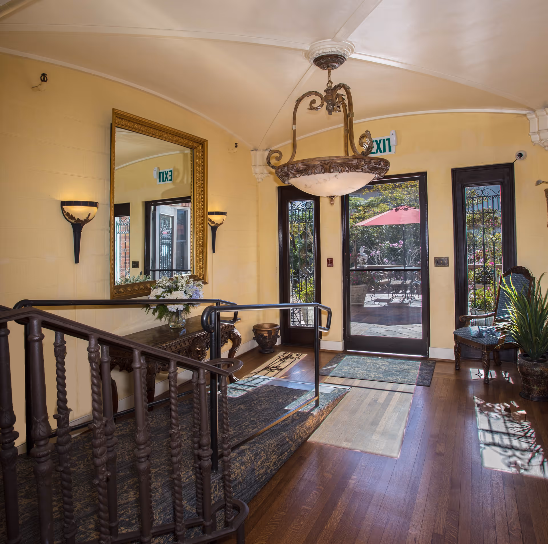 Interior view of an entrance area in a senior living facility with wooden flooring, a decorative wooden railing, a large ornate mirror on the wall, two wall sconces, a chandelier hanging from the ceiling, a small table with a flower arrangement, and glass doors leading outside to a patio with an umbrella and garden.