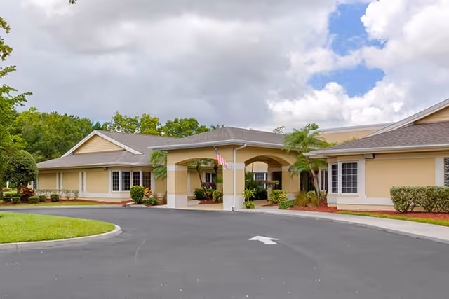 Exterior view of a single-story senior living facility building with beige walls and a covered entrance. The driveway is paved with an arrow pointing forward, and there are landscaped areas with green grass, bushes, and palm trees under a cloudy sky.
