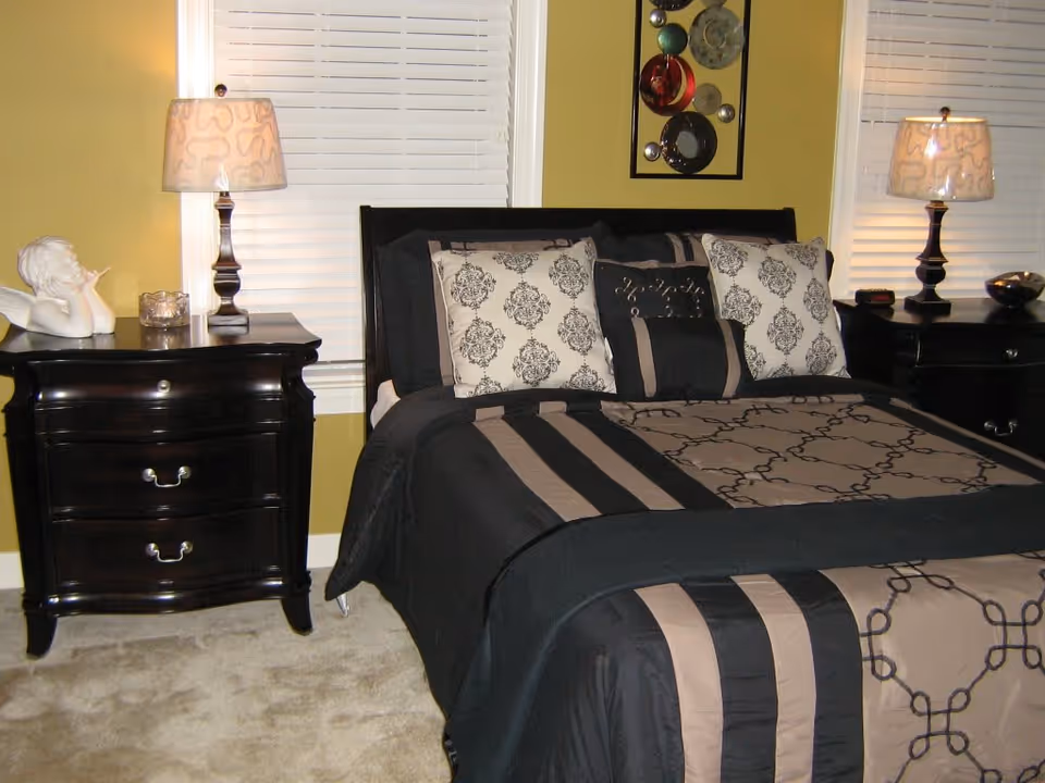 A bedroom with a neatly made bed featuring a black and beige patterned comforter and matching pillows. There are two black nightstands on either side of the bed, each with a lamp and decorative items. The walls are painted yellow, and there are white window blinds behind the bed.