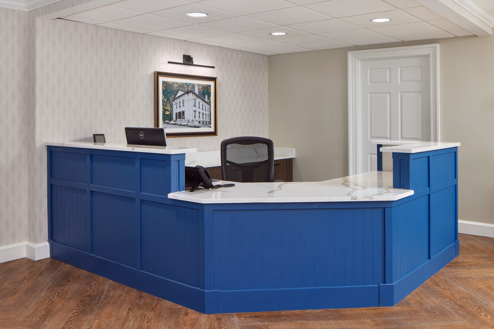 Reception desk area with a blue paneled counter and white marble countertop. Behind the desk is a black office chair, a computer monitor, and a telephone. A framed picture of a building hangs on the wall above the desk, illuminated by a picture light. The room has beige walls, wood flooring, and a white door in the background.
