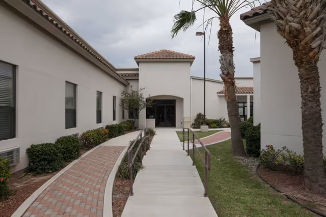 Walkway and ramp leading to the front entrance of a single-story stucco building with palm trees and landscaped beds.