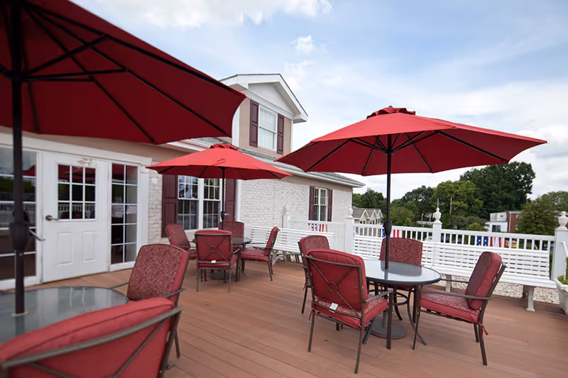 Outdoor patio area with several round tables, each shaded by large red umbrellas. Red cushioned chairs surround the tables. The patio is attached to a white brick building with windows and a door leading inside. Trees and other buildings are visible in the background under a partly cloudy sky.