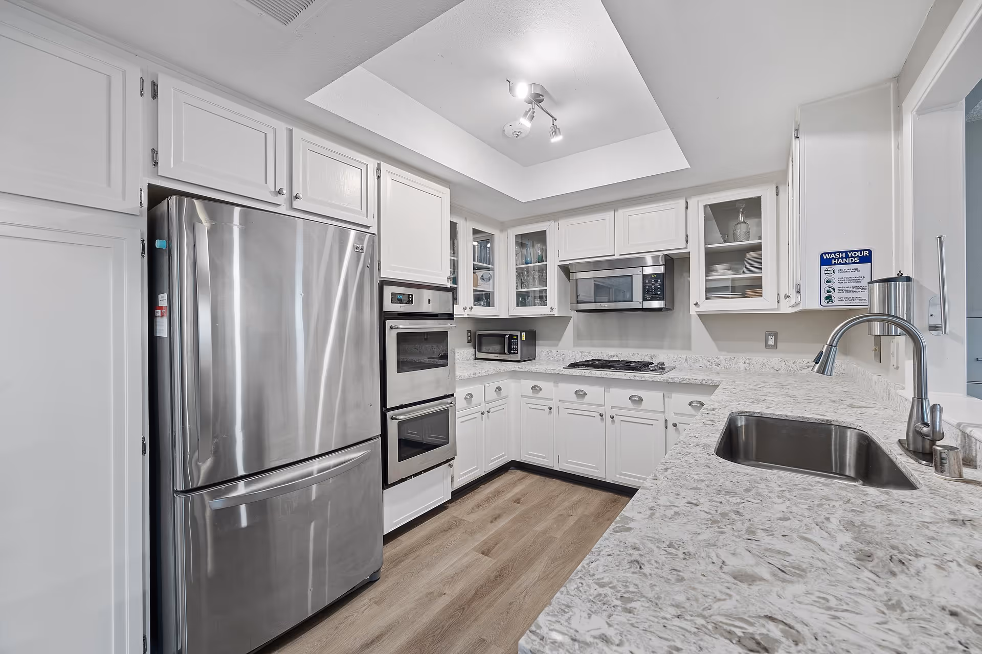 Modern kitchen with white cabinets, stainless steel refrigerator, double oven, microwave, gas stove, and a sink with a faucet. The countertop is light-colored with a marbled pattern. A sign on the wall near the sink reads 'WASH YOUR HANDS' with hygiene instructions.