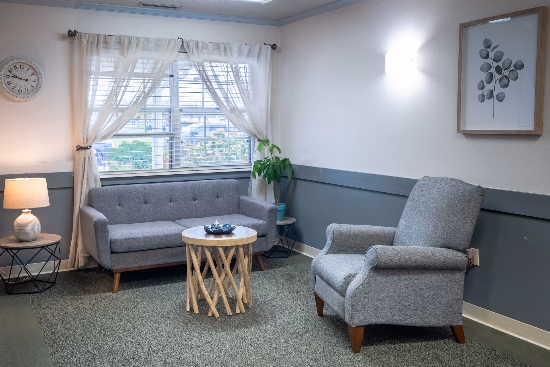 A cozy sitting area with a gray upholstered sofa and matching armchair, a round wooden coffee table with a decorative bowl, a side table with a lamp, a potted plant, a wall clock, and a framed botanical print. Sheer white curtains cover a window letting in natural light.