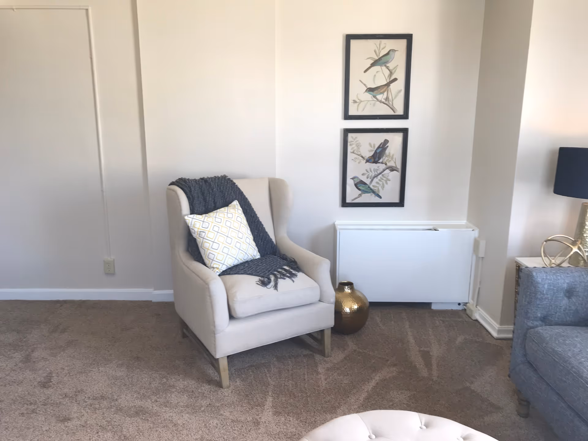 A cozy corner of a living room featuring a beige armchair with a patterned pillow and a dark gray throw blanket. Two framed bird illustrations hang on the wall above a white radiator cover. A gold decorative vase sits on the carpeted floor next to the radiator. Part of a gray sofa and a lamp with a dark shade on a side table are visible on the right side.