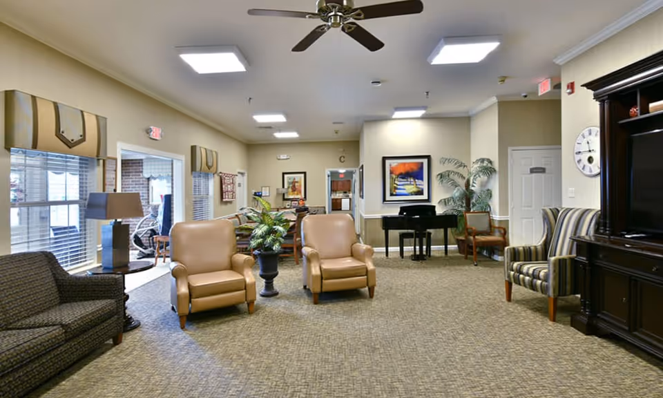 Spacious senior living common room with armchairs, sofa, piano, TV cabinet, and entryway.