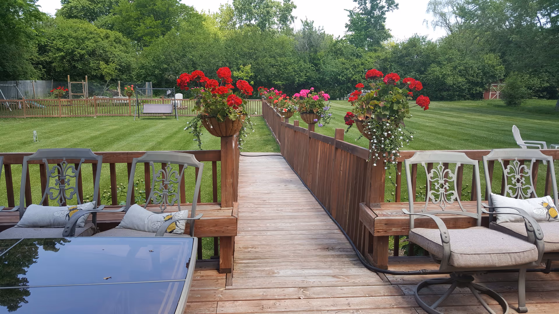 Outdoor wooden deck area with cushioned metal chairs and a glass-top table. The deck has a wooden railing adorned with hanging flower baskets containing red and pink flowers. Beyond the deck is a large, well-maintained grassy lawn bordered by trees and shrubs.