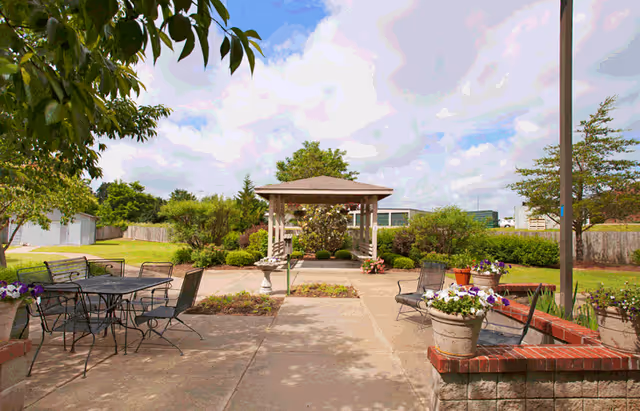 Outdoor patio area with metal tables and chairs, surrounded by flower pots and greenery. A wooden gazebo is visible in the background under a partly cloudy sky.
