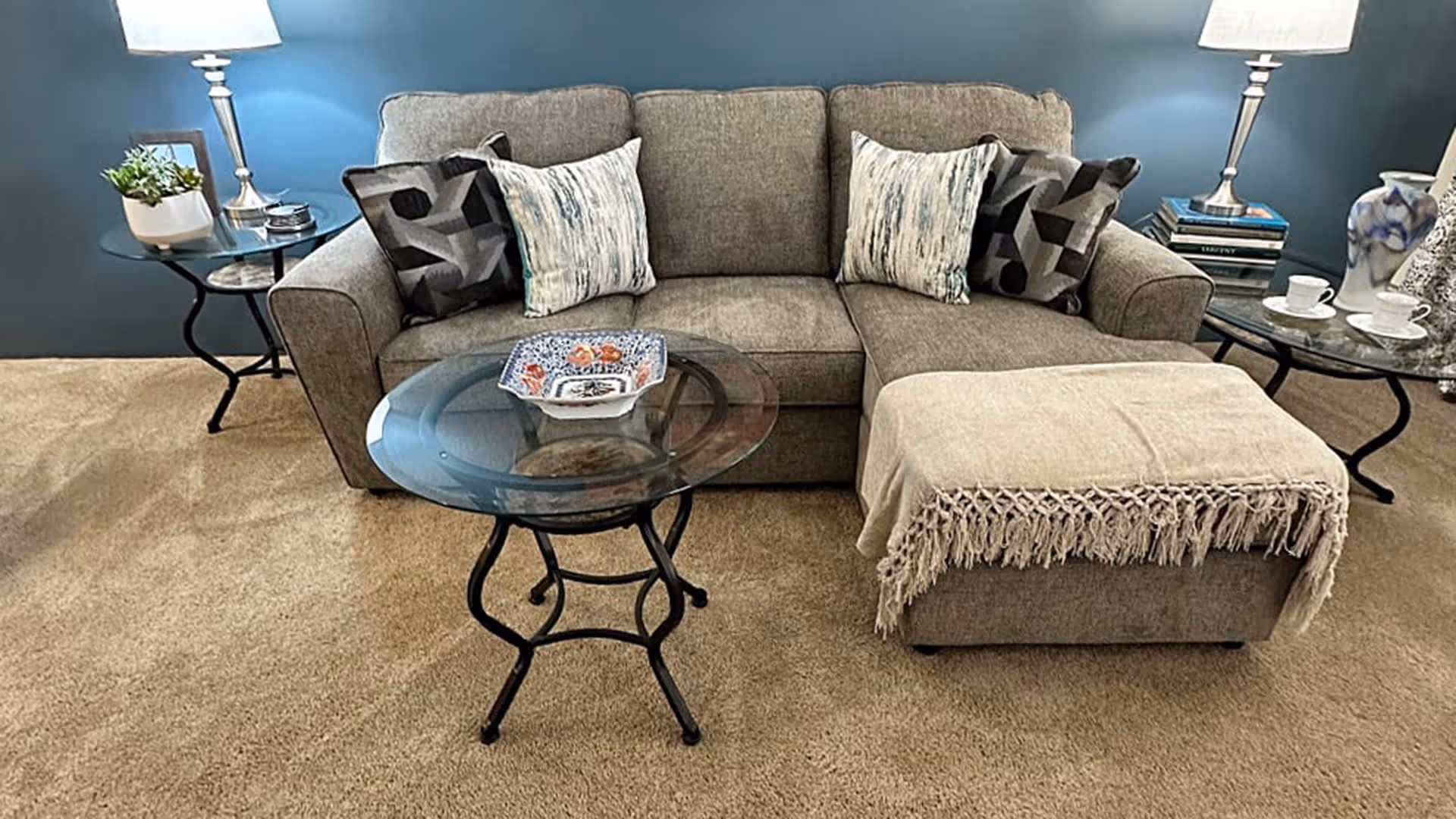A cozy living room area with a gray sectional sofa adorned with patterned and textured throw pillows. In front of the sofa is a round glass coffee table with a decorative dish on top. To the right of the sofa is a matching ottoman draped with a beige fringed throw blanket. On either side of the sofa are small round side tables with lamps, books, a plant, and tea cups. The wall behind the sofa is painted blue and the floor is carpeted in beige.