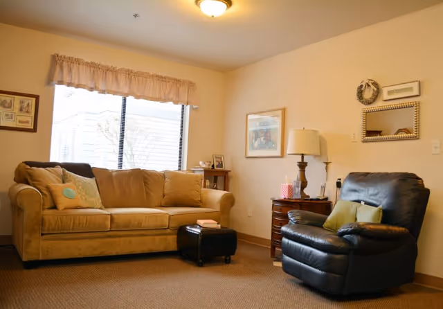 A cozy living room with a beige sofa adorned with multiple cushions, a black leather recliner chair with green pillows, a small wooden side table with a lamp, tissue box, and framed pictures on the wall. A window with a beige valance lets in natural light.