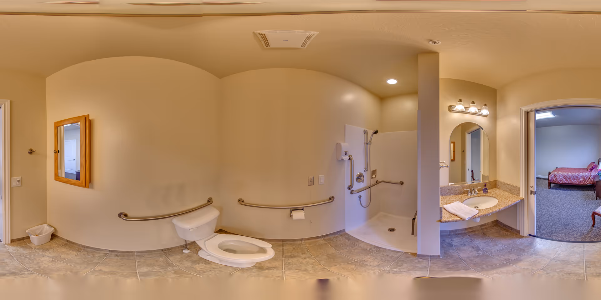 A spacious bathroom with beige walls and tiled floor, featuring a toilet with grab bars, an open shower with grab bars and a handheld showerhead, a sink with a granite countertop and a mirror above it, and a doorway leading to a bedroom with a bed and chair visible.