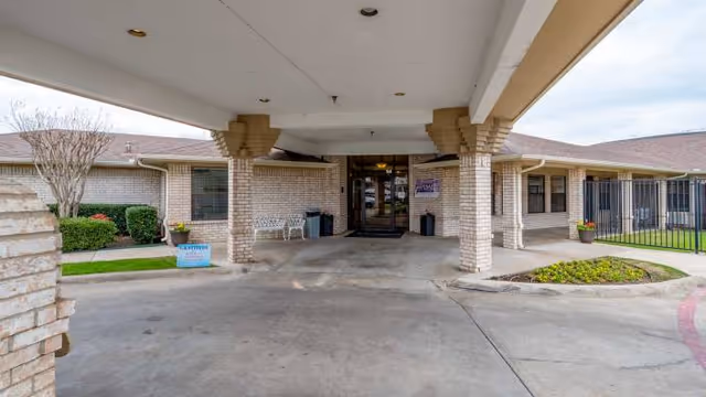 Entrance area of a single-story brick building with a covered driveway supported by columns. There are bushes, a small tree, and flower pots near the entrance. A bench and trash bins are visible near the door.