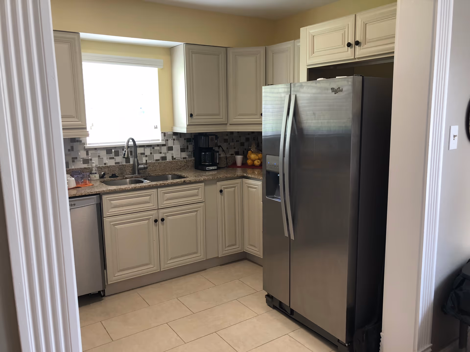 Compact kitchen with cream cabinets, a double sink under a window, tiled backsplash and a stainless steel side-by-side refrigerator.