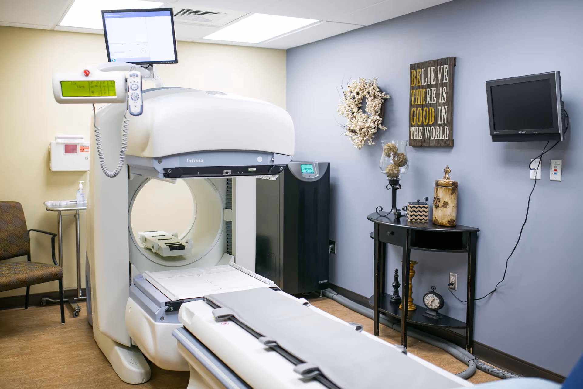 A medical imaging room featuring a large white imaging machine with a patient bed, a monitor mounted on top, and a control panel. The room has light-colored walls, a small table with decorative items including a wreath, a clock, and a wall-mounted TV. There is also a chair and a small side table with hand sanitizer in the corner.